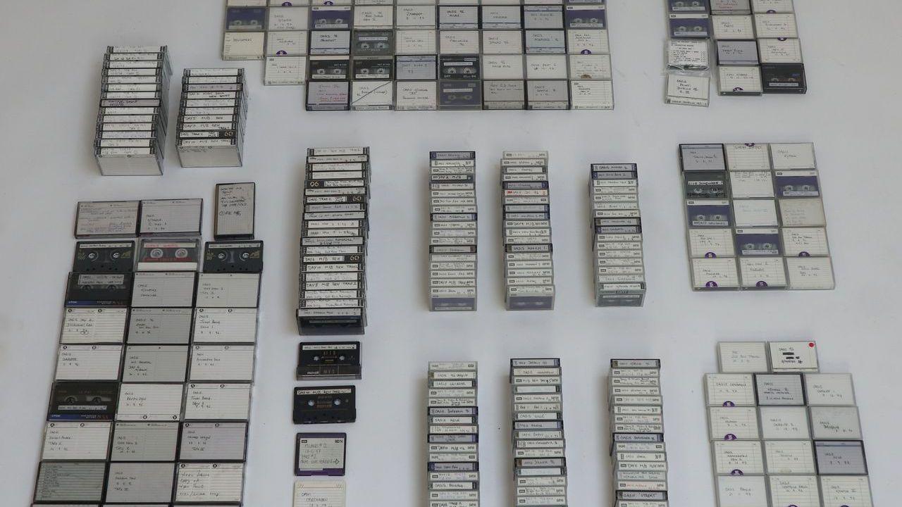 Dozens of tapes are grouped together and laid out on a white surface. There is small hand written notes on each tape.