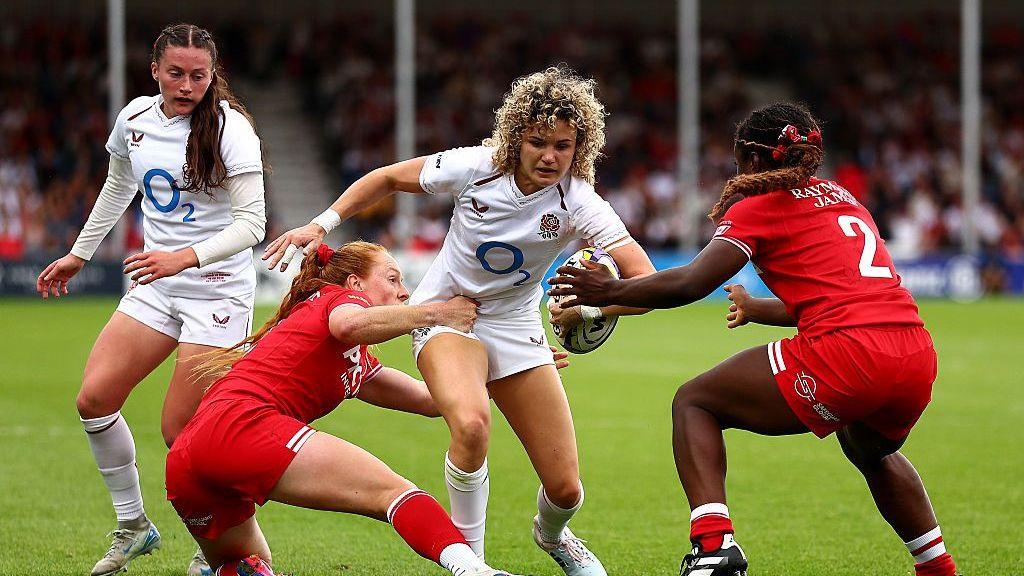 An England player, wearing white, is tackled by two Canada players in red