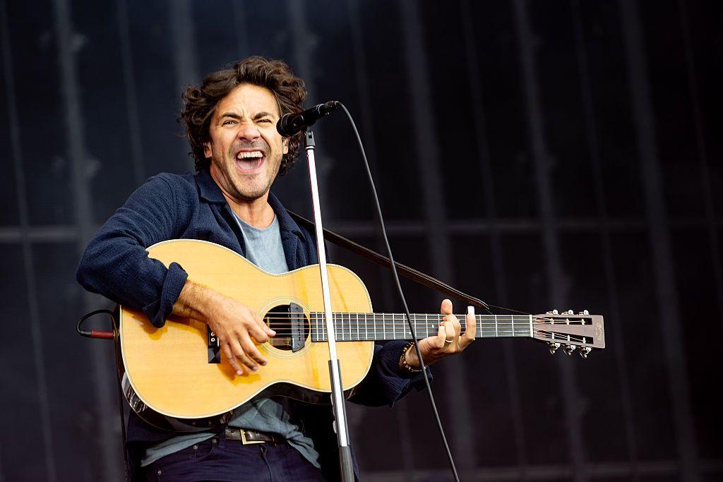 Jack Savoretti performing on stage. He is mid song and is playing an acoustic guitar. He has dark, floppy hair and is wearing a grey T-shirt and a dark shirt or jacket over the top.