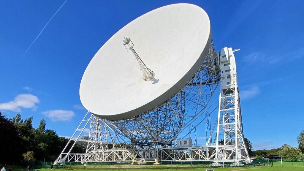 A huge white radio telescope aligned with its large dish facing the sky, as seen from tree cover.