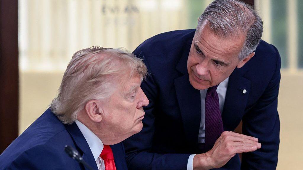 A file image showing U.S. President Donald Trump at a working lunch with Prime Minister Mark Carney and other G7 leaders. The image shows Carney leaning over Trump while the two are mid-conversation. Both are wearing a suit. Carney is gesturing with his hand mid-speech, while Trump appears to listen with a neutral expression on his face..