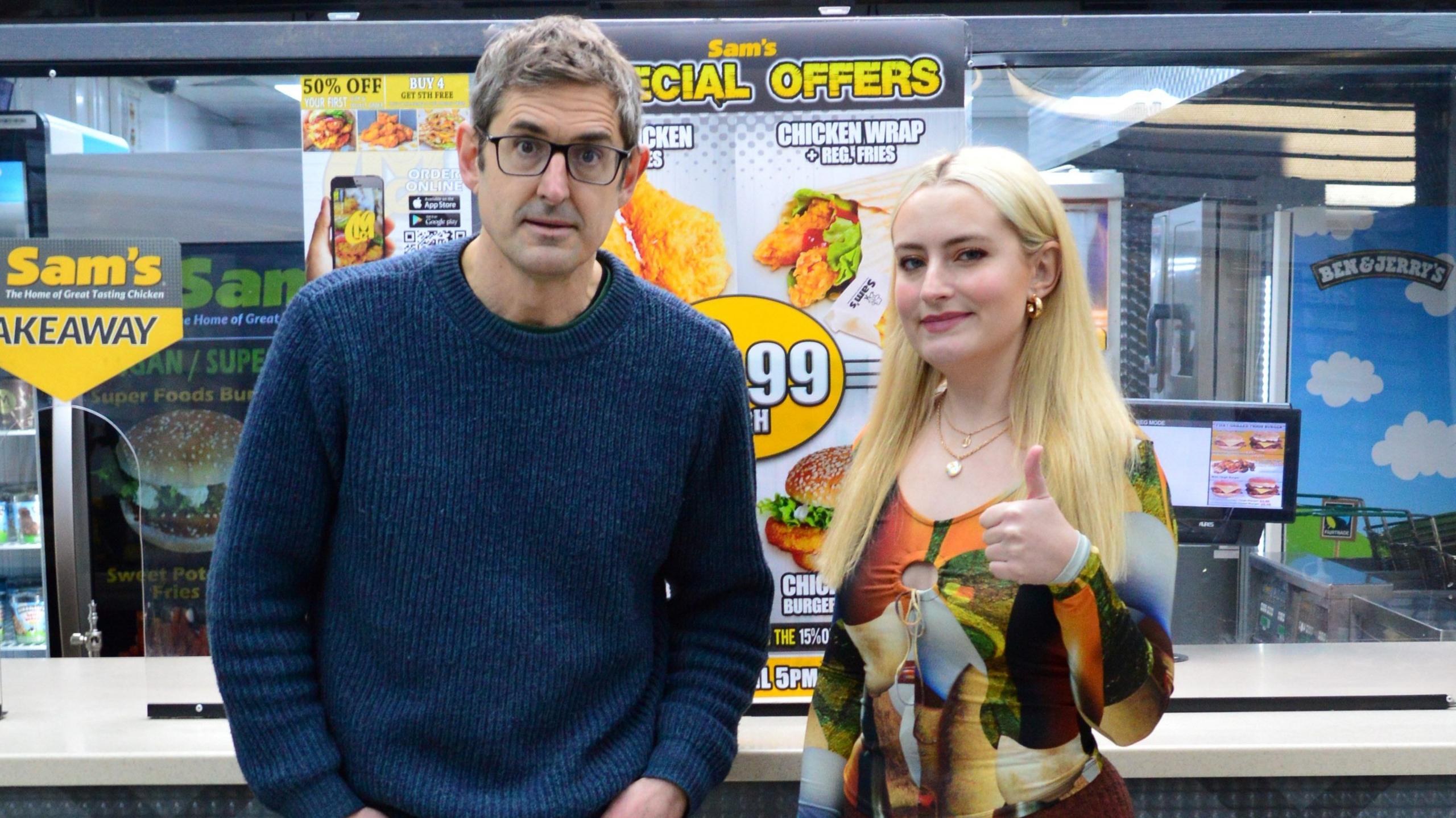 Louis Theroux and Dimoldenberg pictured at a counter in a chicken shop. Theroux, in his 40s, has greying hair and dark rimmed glasses. He wears a blue cable knit jumper and poses with his hands in his pockets. Dimoldenberg, wearing a patterned top, gives the camera a thumbs up.