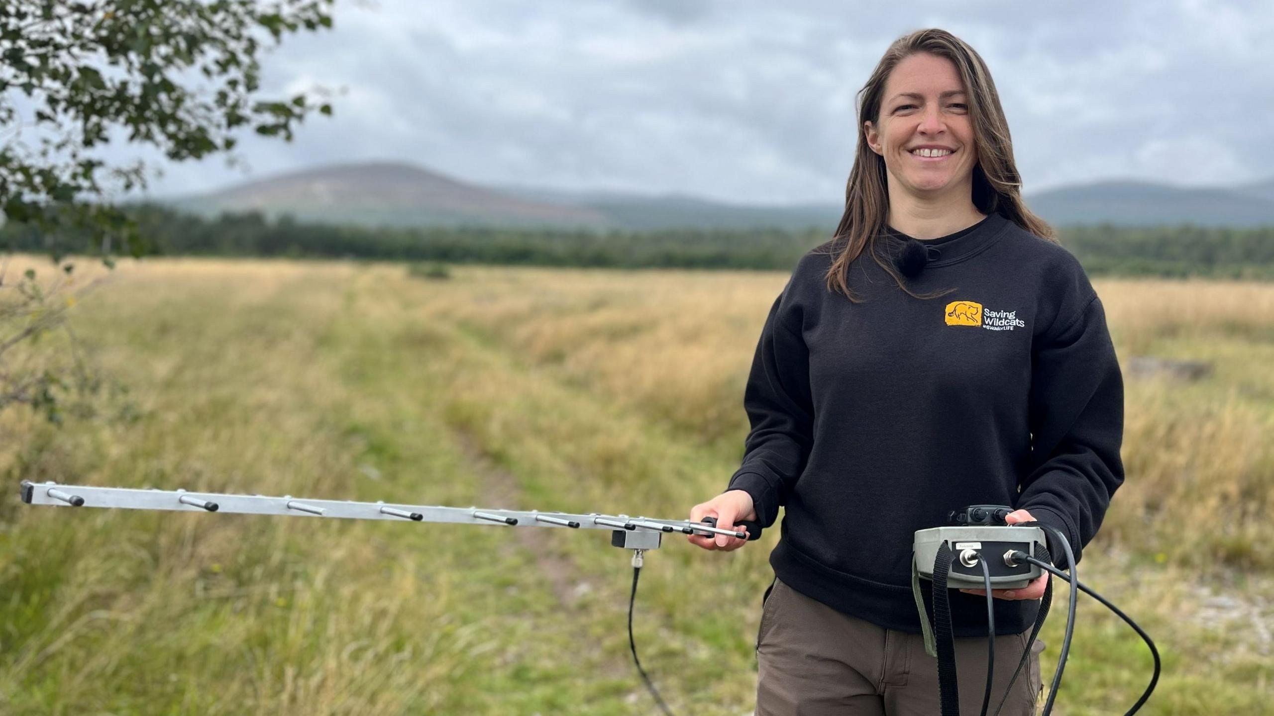 A young woman with long brown hair stands in a field, holding a monitoring device on both hands. She is wearing a dark sweatshirt which says "Saving wildcats".