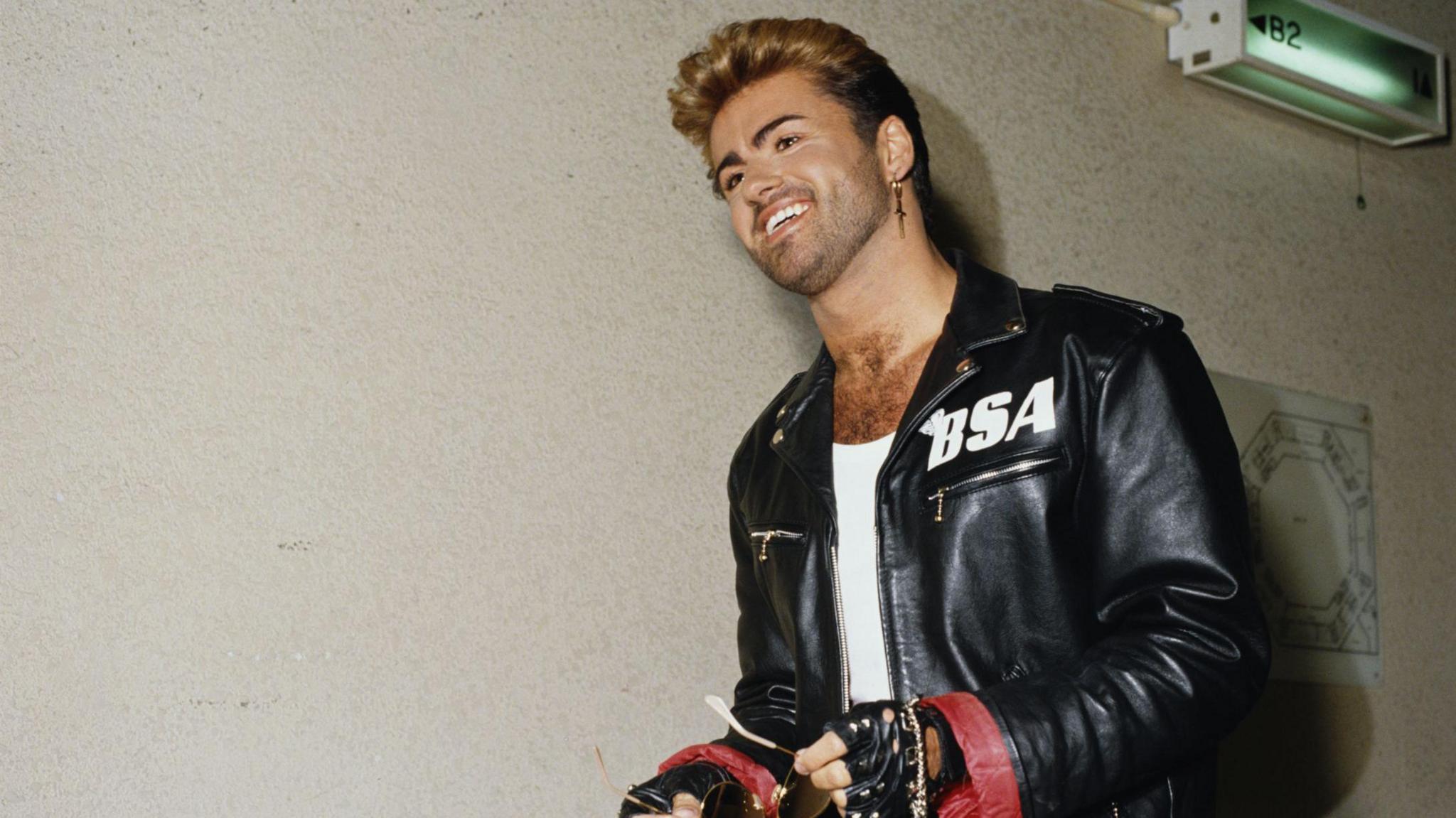 George Michael wearing a black jacket with a white BSA logo on the left side.