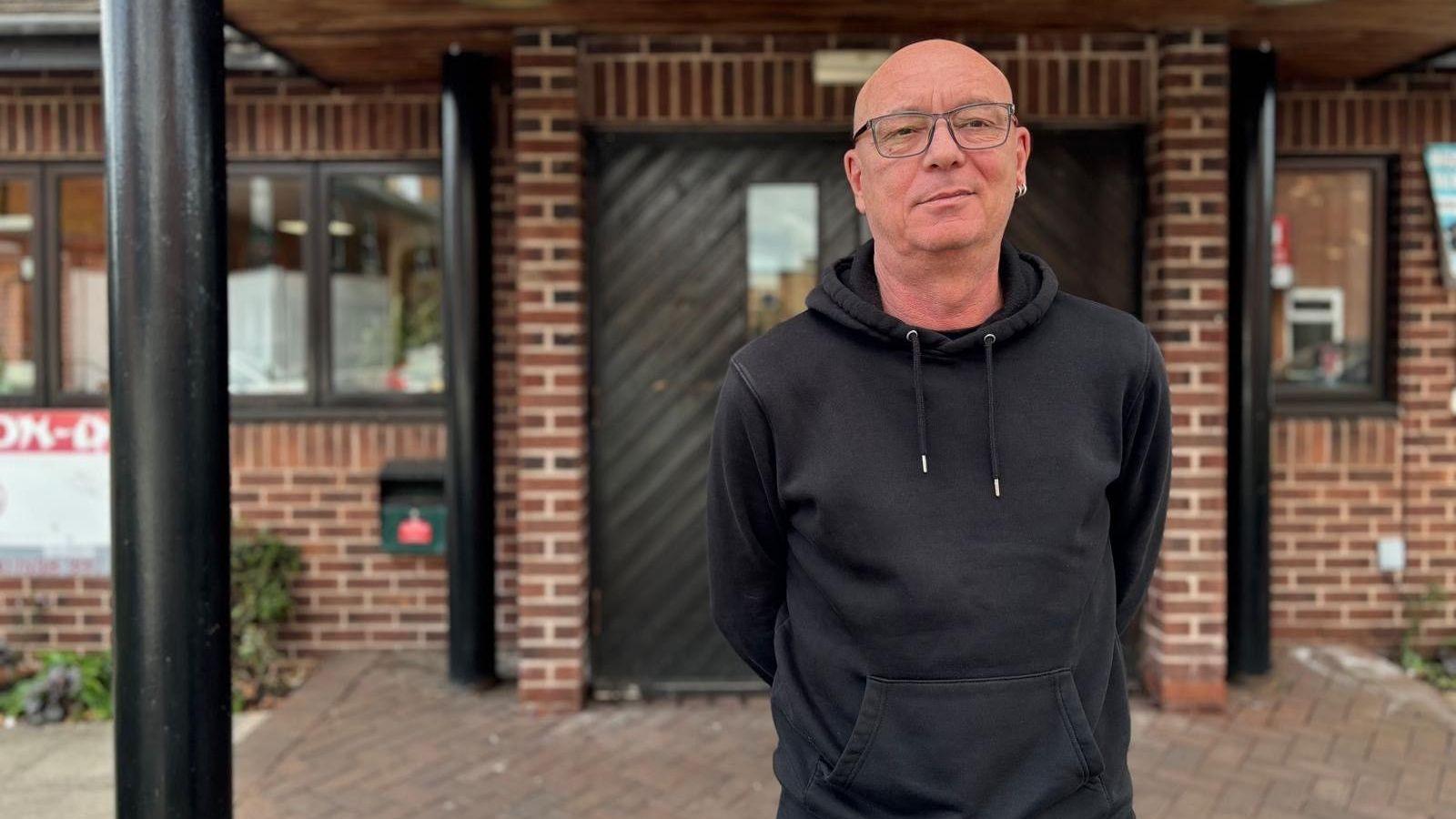 A man in a black hoodie and black rimmed glasses stands with his arms behind his back. He is in front of a brown brick building.