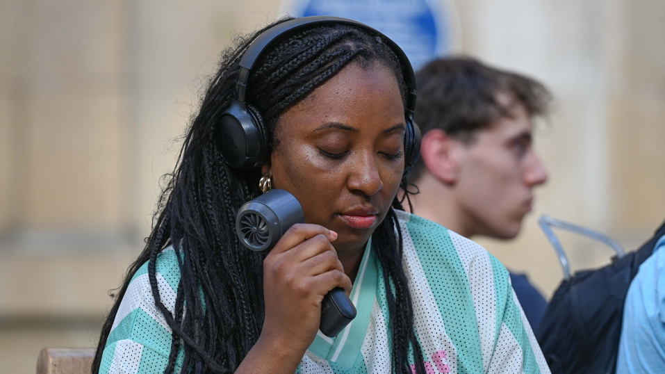 A woman in a green and white striped shirt holds an electric fan to her face during a heatwave