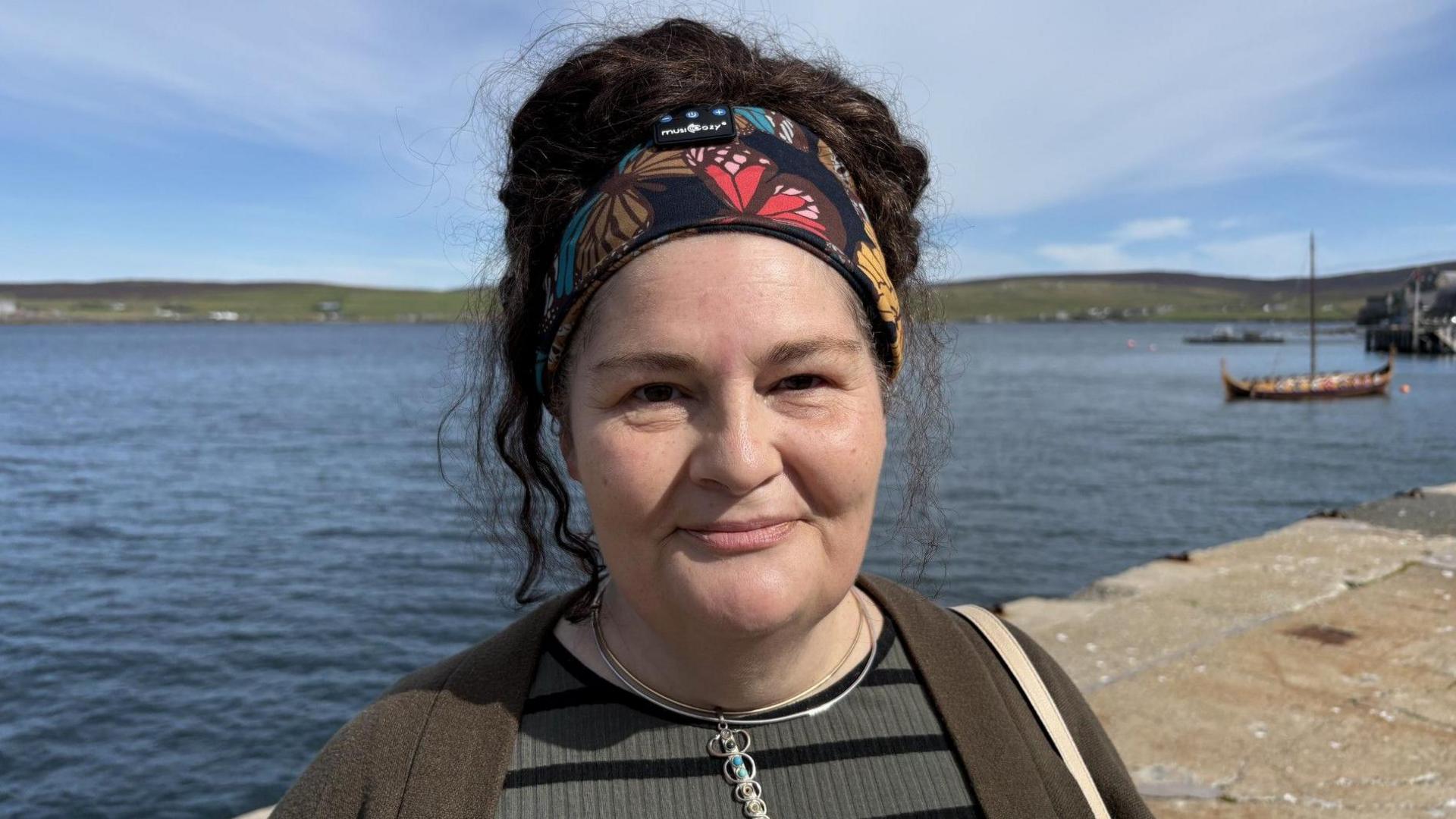 Diane Barnes is a 51-year-old woman with black hair. She is looking at the camera and standing in a harbour area. Sea and hills are behind her and there's a small viking boat to the right. She is wearing a green top, necklace and her her is held up by a bandanna.