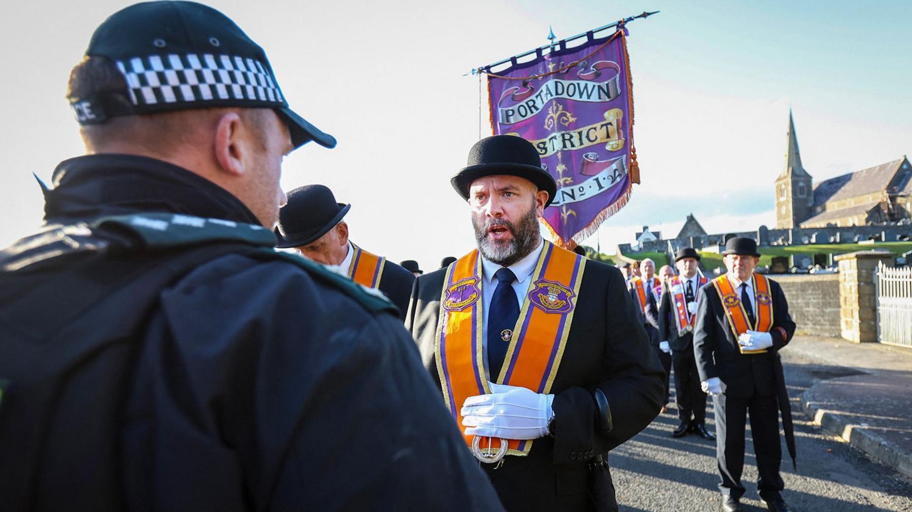 An Orange Order member talking to a police officer wearing a black suit, black hat and an orange sash. The rest of the lodge members are behind him with the church in the background. The police man is wearing a black and white checked hat with a black coat with green PSNI lapels.