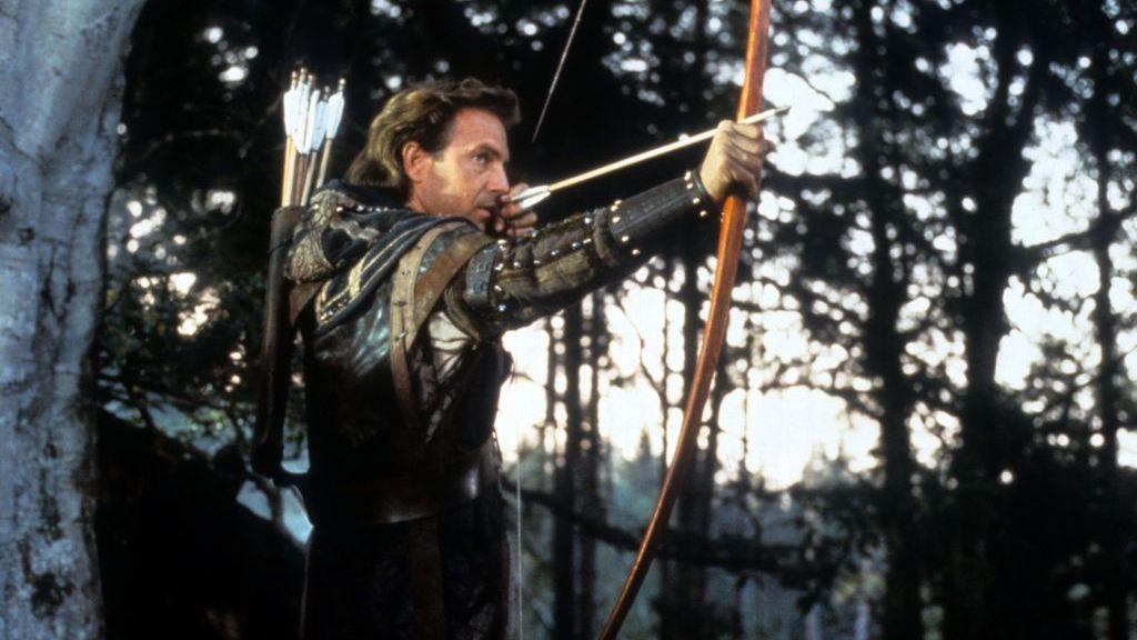 A man with his back to a white tree prepares to fire a wooden bow, with an arrow loaded in it. He wears a black and brown leather suit. He is in a wooded area.