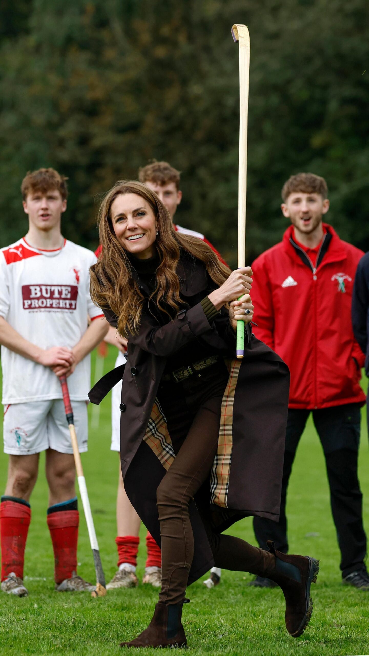 The Princess of Wales dressed in a brown coat, top and jeans swingsa curved wooden stick called a caman during a game of shinty, as three men dressed in red and white sports gear watch on.