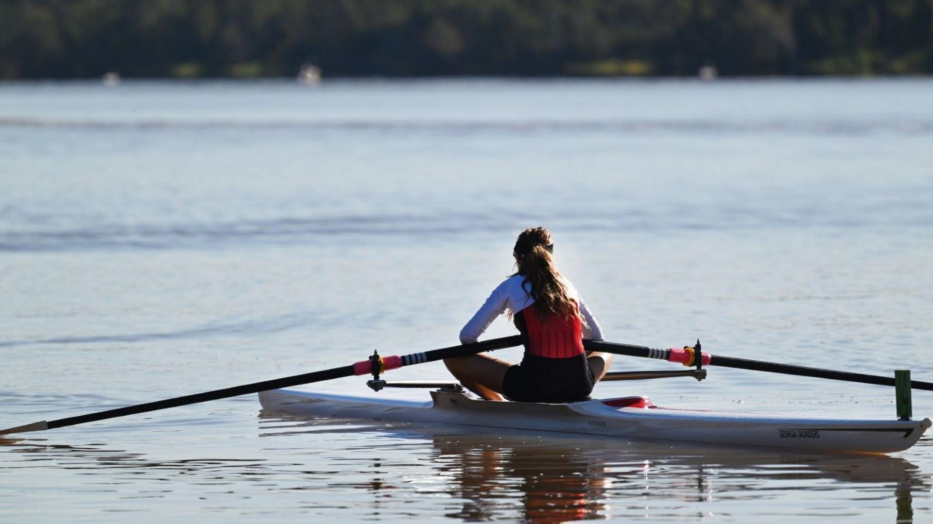 A girl in rowing wear sits in a scull on a wide river with her back to the camera