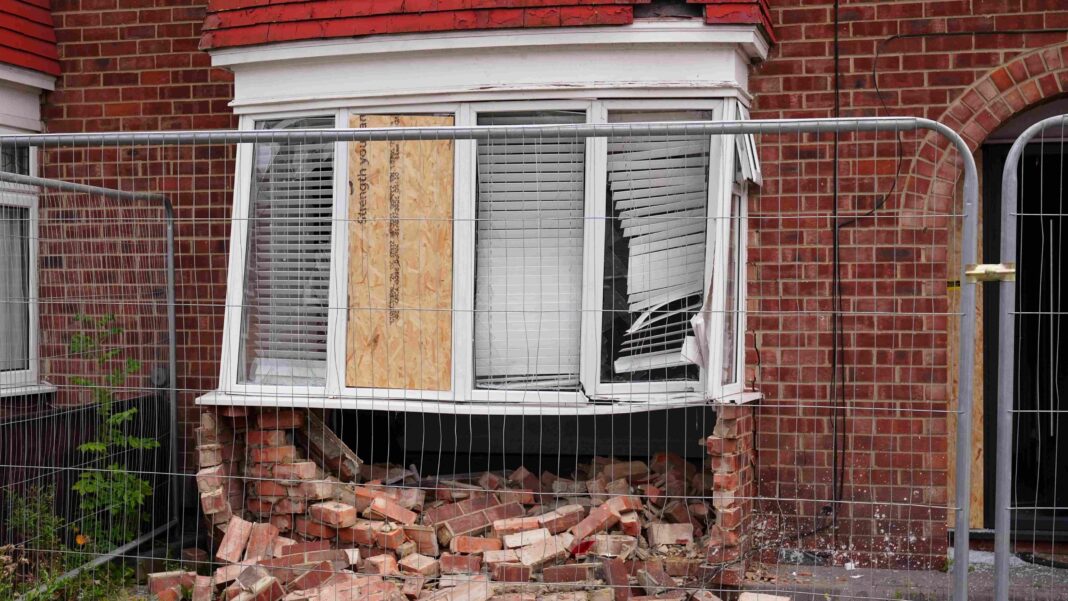 The badly damaged exterior of a brick home. The front window has been smashed, with one pane boarded up, and the bricks underneath have collapsed. Metal fencing has been put up around the area.