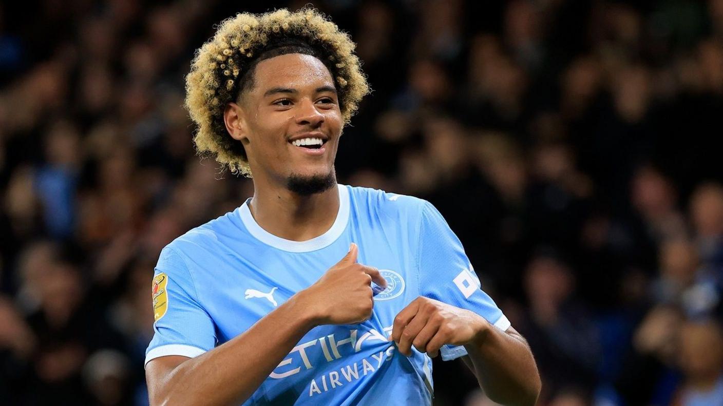 Floyd Samba smiles while pointing to the badge on his Manchester City shirt