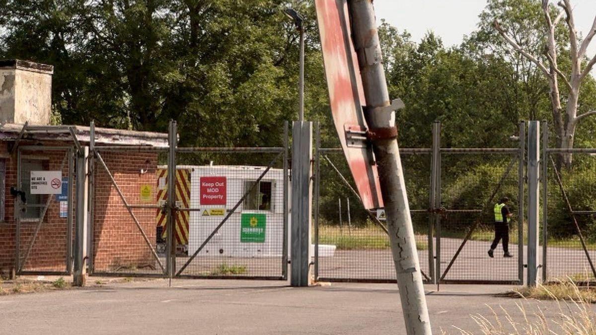 The gates outside MoD Bicester site A. A security guard is patrolling behind gates and a barbed wire fence.