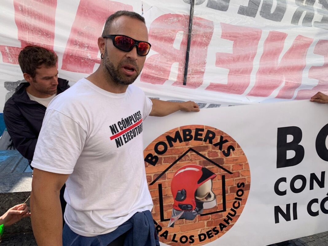 Diego Blanco stands holding part of a big sign representing firefighters at the Madrid housing protest. He has short dark hair, and is wearing sunglasses and a white T-shirt.