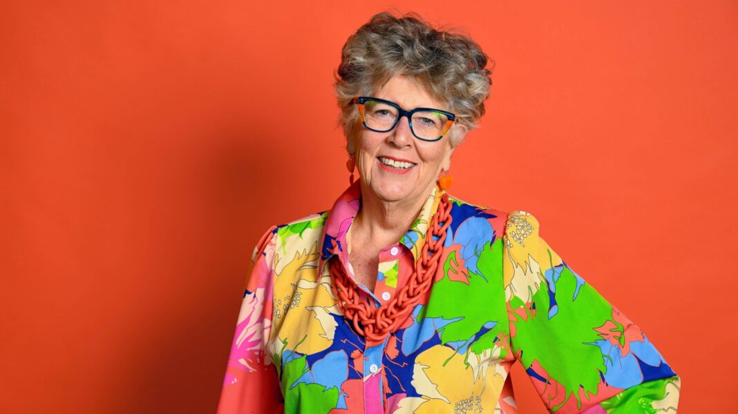 Prue Leith: ‘You come into your own growing old’ Dame Prue Leith smiles as she poses with her hand on her hip in front of a bright orange background. She is wearing very colourful glasses and a very colourful floral blouse, along with an oversized orange chain necklace. She has short grey hair.