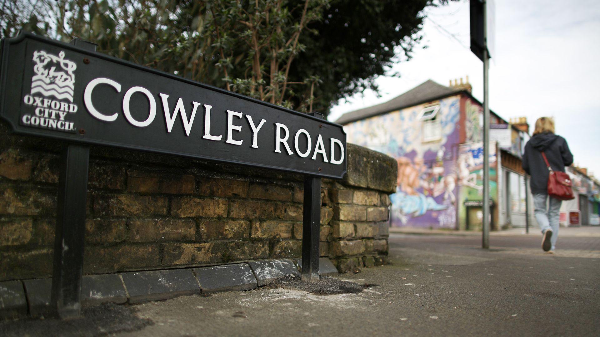 A street-name sign with white letters against a black background reads "Cowley Road", with the words "Oxford City Council" to the left, in a smaller size. A woman with a red shoulder bag, and wearing a black coat and blue jeans, is walking away into the distance. There is colourful graffiti on the side of the building in the distance.