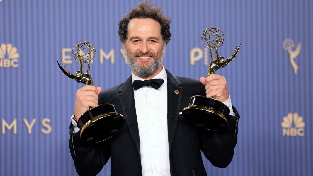 Matthew Rhys is photographed attending the 78th Emmy Awards. He is wearing a black suit jacket, black waist coat and black bow tie. He has dark hair and a grey beard. He is looking at the camera and smiling whilst holding two Emmy awards.