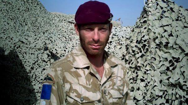 A profile image of Keith J Abraham in his army uniform, a burgundy beret and a camouflage shirt. Behind him is camouflage netting.