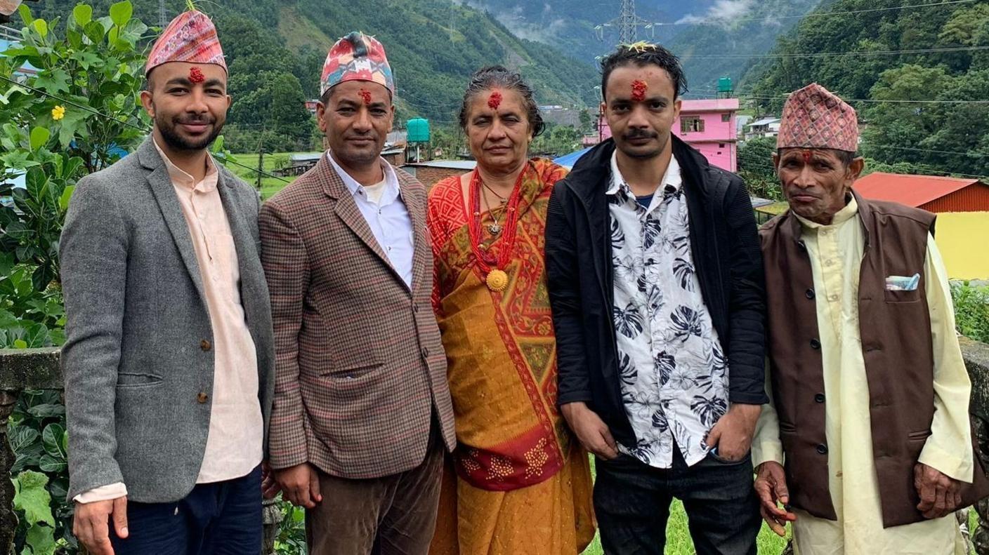 Five people stand outdoors in front of hills and buildings in a rural area. Trees and mountains are visible in the background.