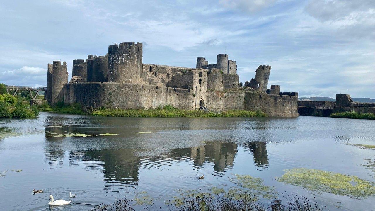 The ruins of Caerphilly castle sit in by a lake with grey, blue sky reflected in the water