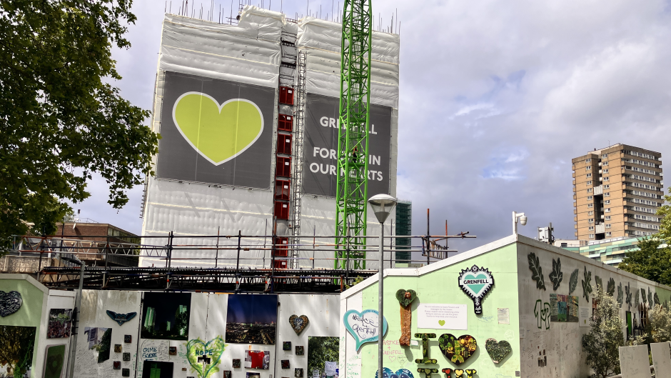 A partially demolished Grenfell tower with a wall surrounding the area which is covered in tributes and drawings. The building is covered in a wrap which features a green heart.