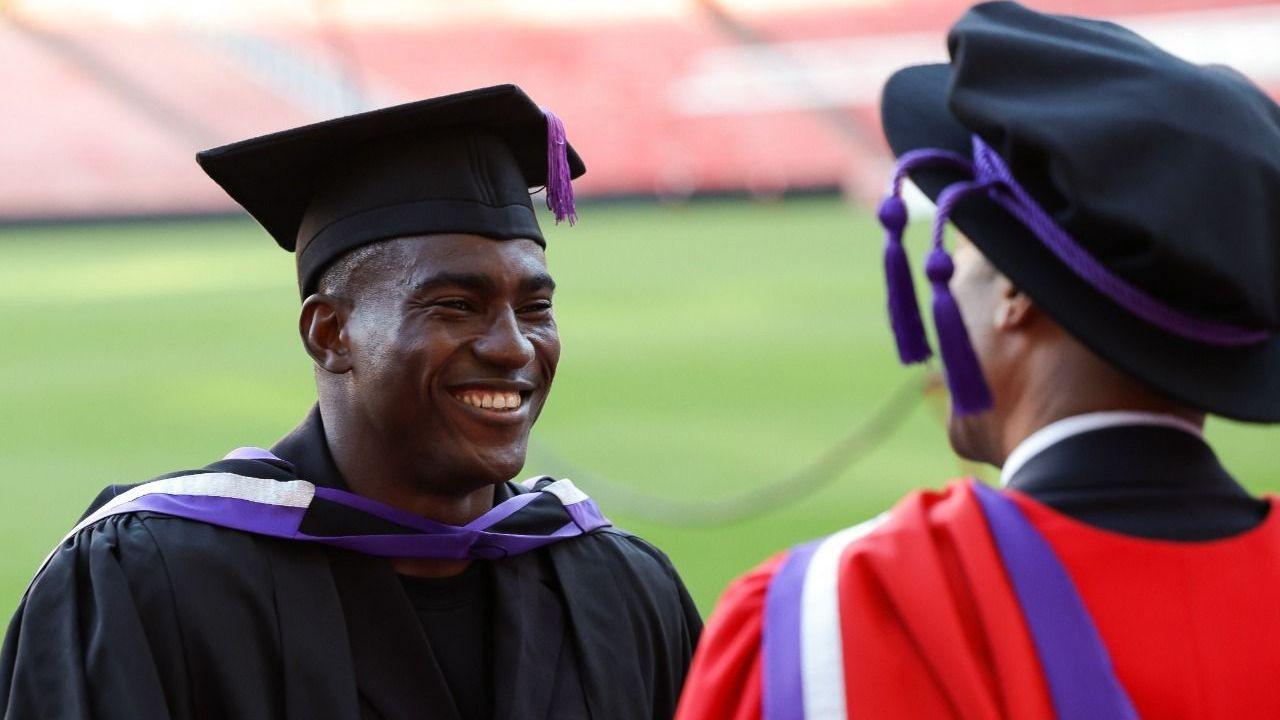 Taiwo Awoniyi smiles as he wears a black graduation gown and mortarboard, with a white and purple stole across his shoulders