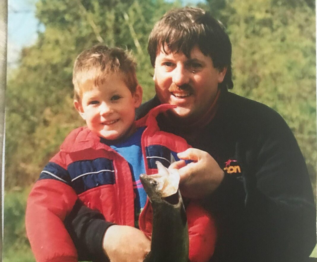 Brett is on the left and a young boy in this undated photo. He is wearing a red jacket and he's smiling at the camera. He is in the arms of his dad Bruce who is wearing a black jacket and is holding a fish in his left hand.