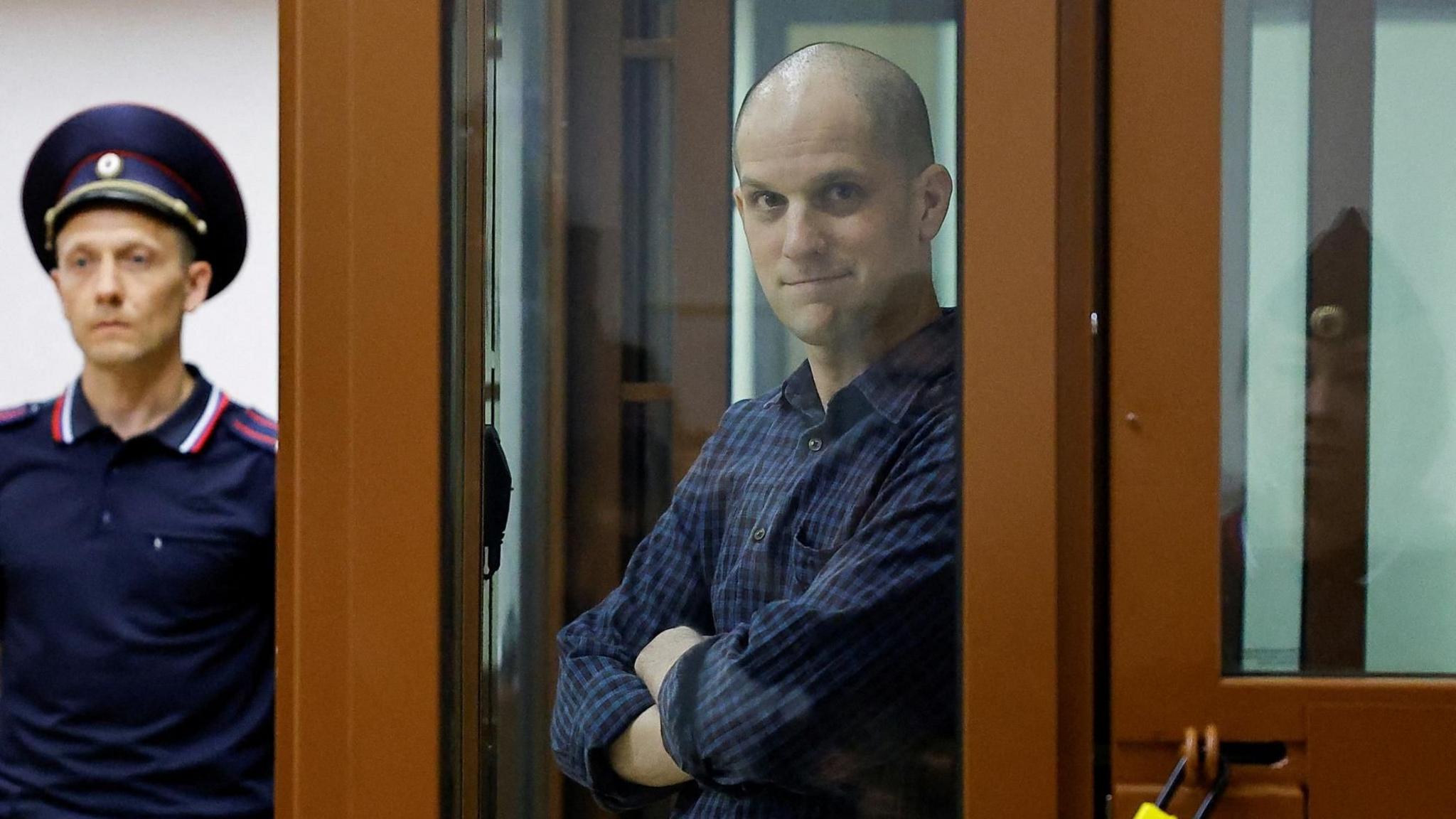 Evan Gershkovich has a shaved head and is wearing a blue checked shirt as he stands behind a glass screen in court. There's a security guard in the background.