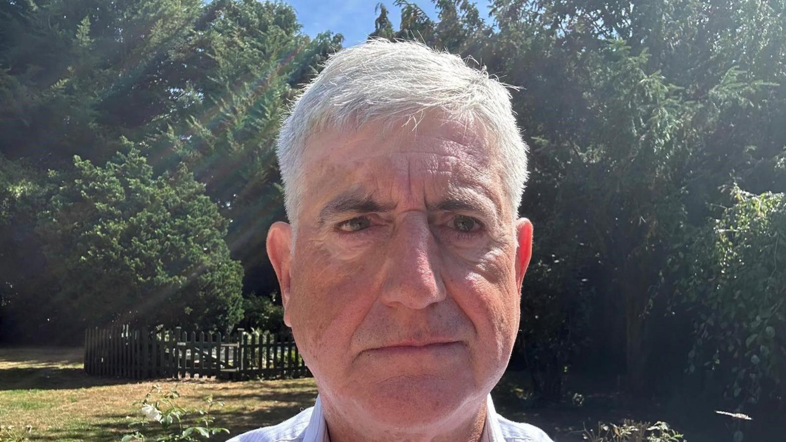Tony Smith is standing in a park with trees directly behind. He is looking at the camera and has short grey hair.