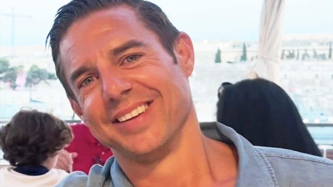 Close-up headshot of a smiling Nicholas Brandram with short dark hair, blue eyes, and clean-shaven facial hair. He is wearing a blue open-collared shirt outdoors, with a blurred background showing an outdoor seating area and buildings under a clear sky.