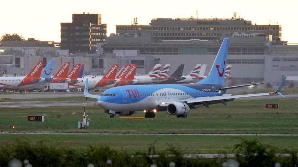 Planes on a runway. Some have easyjet tails while one in the foreground is Tui. The airport is in the background.