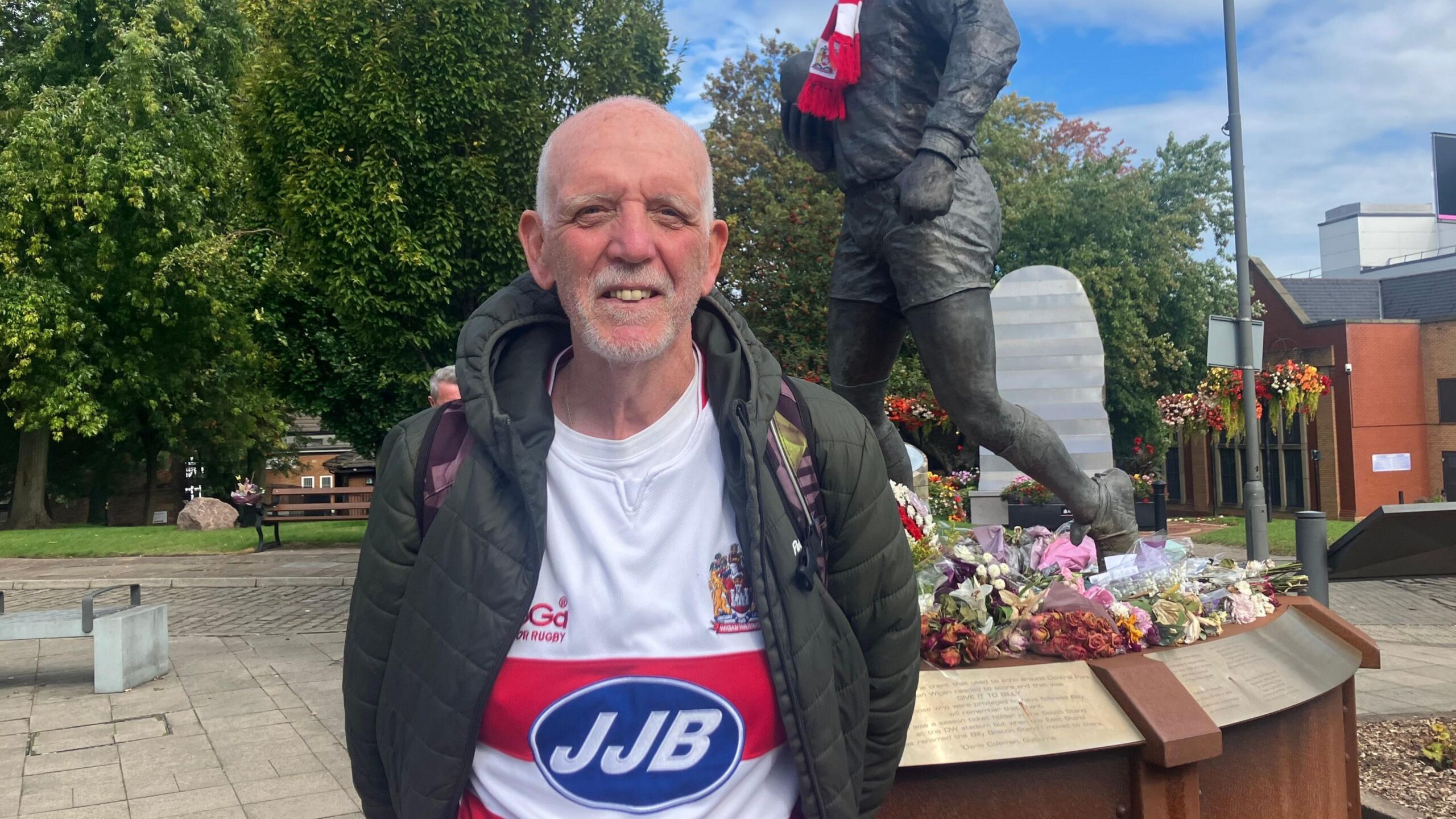 A bald man smiles to the camera. He is wearng a coat and Wigan jersey. Behind him is a statue of Billy Boston holding a rugby ball. He had a red and white scarf around his neck. Flowers surround the base.