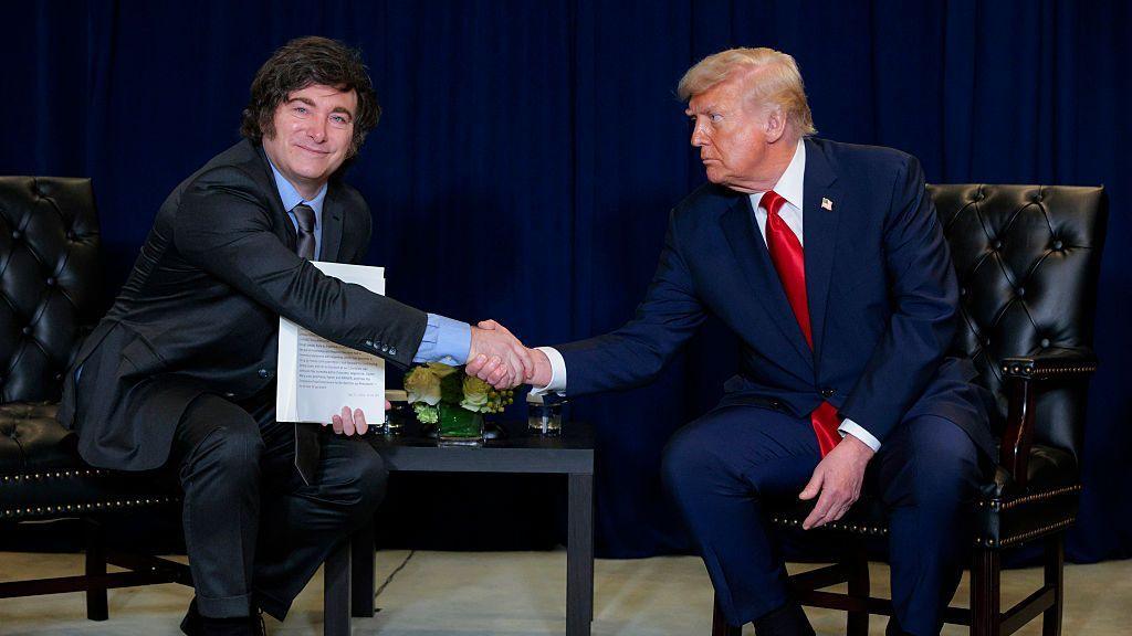 US President Donald Trump (R) shakes hands with President of Argentina Javier Milei during a bilateral meeting at the 80th session of the UN’s General Assembly at the United Nations headquarters on September 23, 2025 in New York City. The men are both wearing dark suits and sitting on chairs. Milei is smiling with his mouth closed, while Trump, looking at him, has a serious expression.