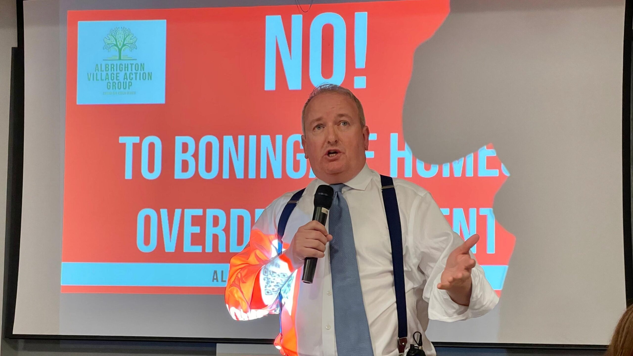MP Mark Pritchard wearing a blue tie, which shirt and braces, standing in front of a projector screen showing a red sign with the words "No to Boningale Homes overdevelopment". He is holding a microphone.