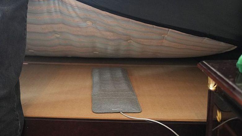 A slim rectangular grey pad with a white wire coming out the end is laid on a wooded board and you can see a mattress being held up above it.