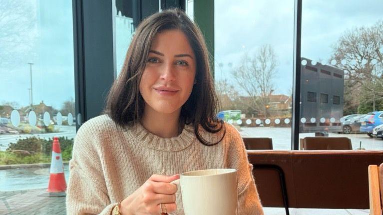 A close up image of Erin Atkinson wearing a cream jumper and holding a mug. She appears to be sitting in a public space with a car park seen out the window behind her.