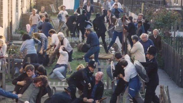 A screenshot from the Our Friends in the North scene where there is a violent disturbance in a pit village. It looks to be a mass fight with the police in the front gardens of terraced houses. Fences have been knocked over and there are multiple people on the ground. There are people of all ages there.