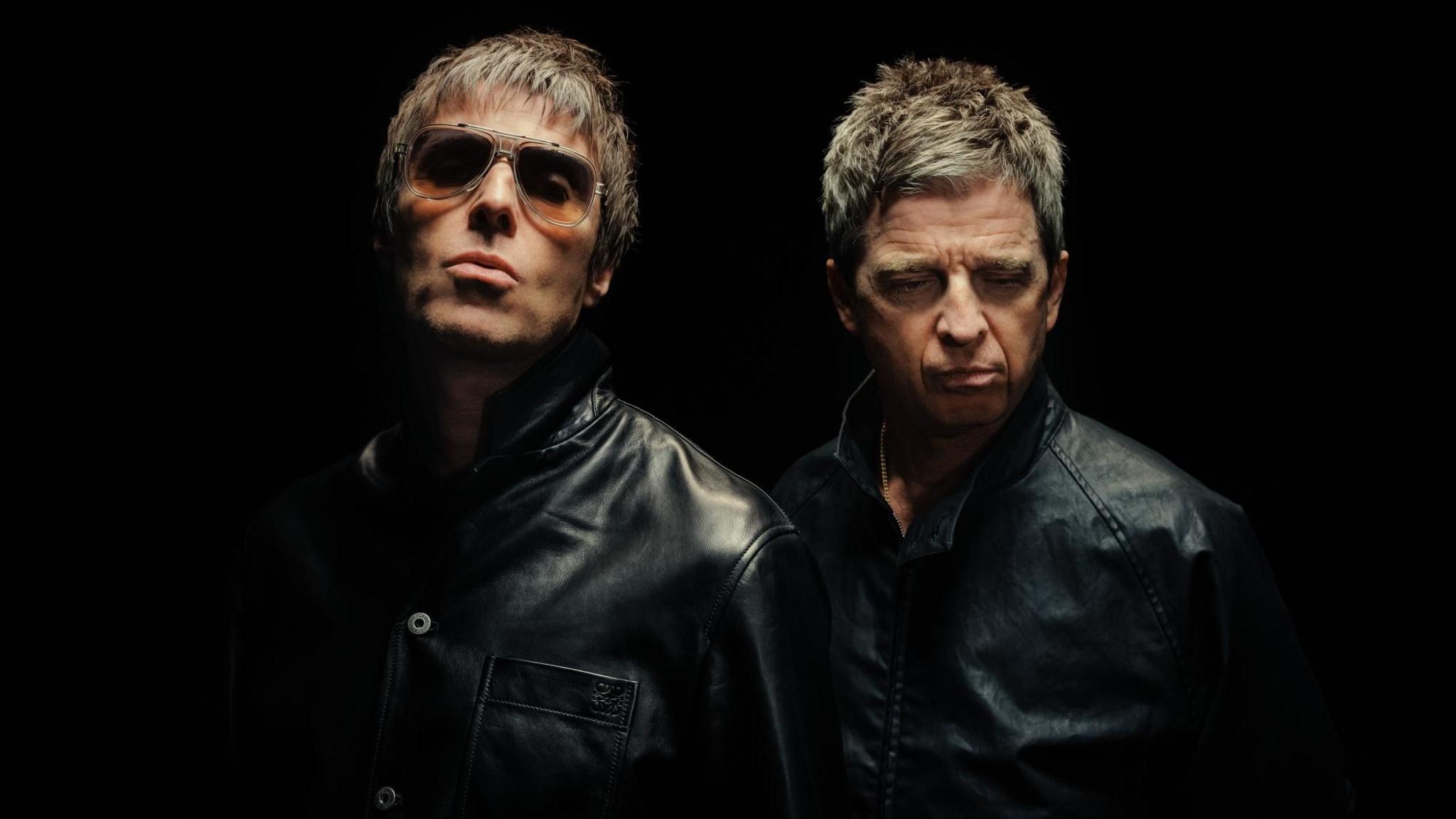 A recent photo of Liam and Noel Gallagher stood together wearing black leather jackets, partially obscured by shadows, in a dark room. Liam is wearing shades and looking at the camera, while Noel is looking towards the ground.