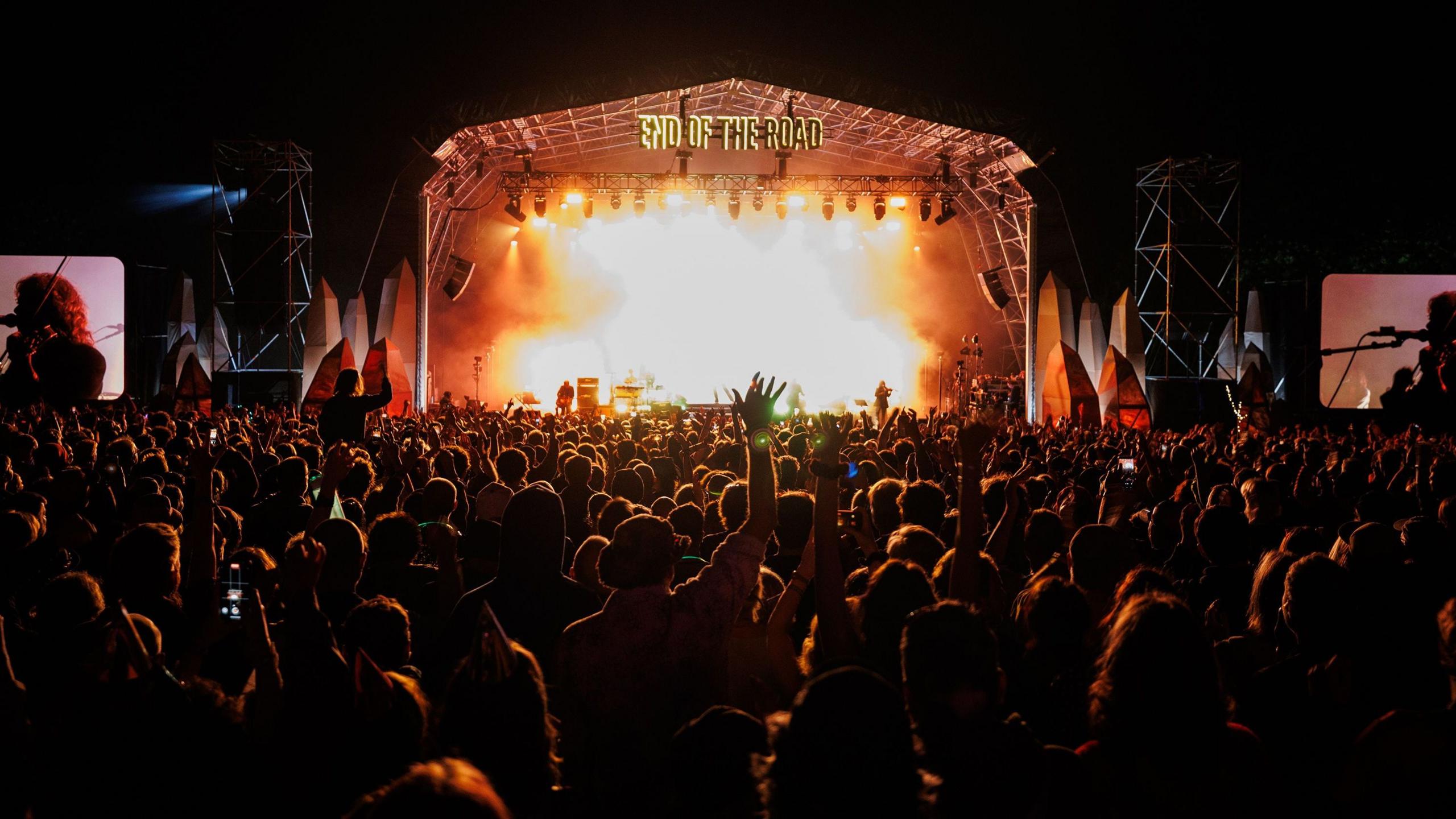 A dark crowd looking at a stage with flames on the stage and bright lights.