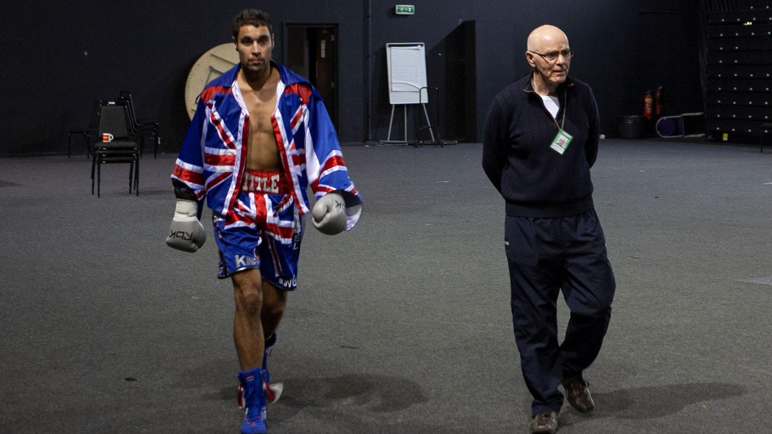 A man in a pair of shorts and a jacket emblazoned with the Great Britain logo walks alongside an elder man with no hair through a backstage area.