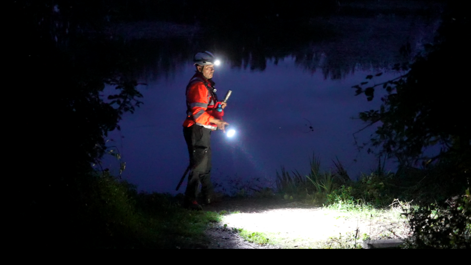 A member of a search and rescue team wearing a hi-vis jacket. He is using a torch and headtorch to search an area next to a pond.