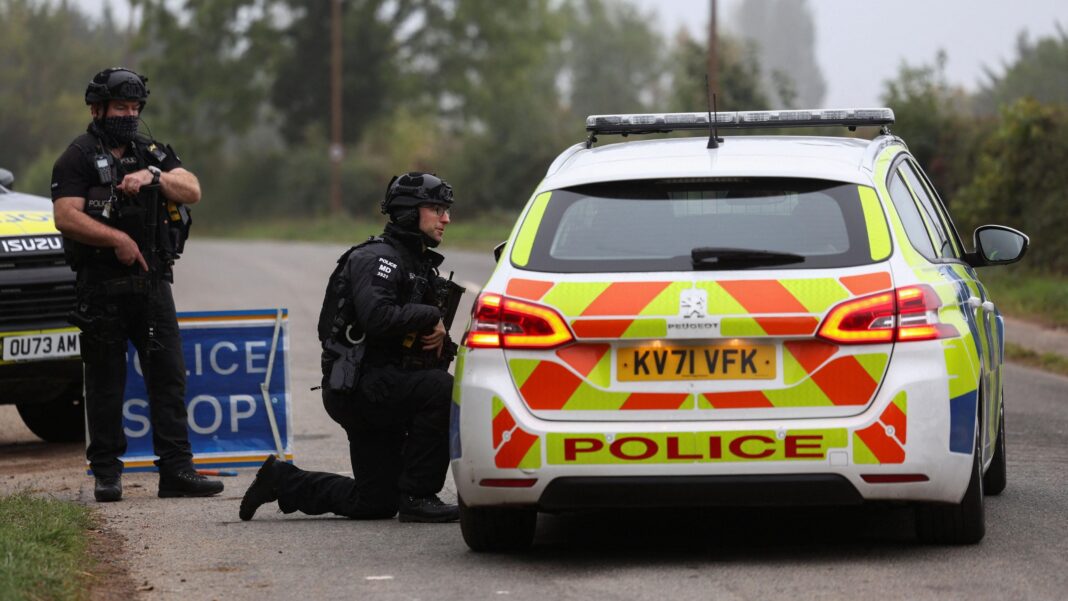 Police officers on 29 September 2026 guard an area when roadblocks were in place at the time near RAF Fairford airbase. The two police officers are carrying guns and wearing firearms uniforms. One officer is standing, the other is kneeling and facing into the window of a police car.