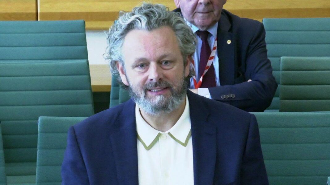 A person with short, curly grey hair and a grey beard sits facing the camera at a desk in a meeting room. They have on a navy jacket over a white shirt with pale green edging around the collar. Rows of empty green upholstered chairs and light wooden panelling are in the background with another person sitting behind.