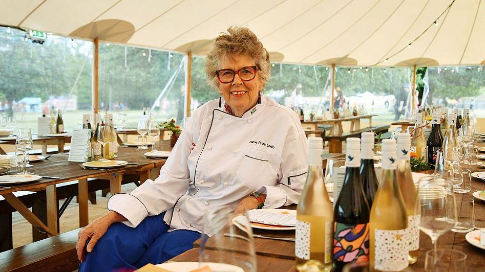 Dame Prue Leith, who is wearing a white chef's shirt and blue trousers, sits down at a long table in the dining area of a festival. She is smiling and has short hair and glasses. There are bottles of wine, wine glasses, and crockery on the tables surrounding her.