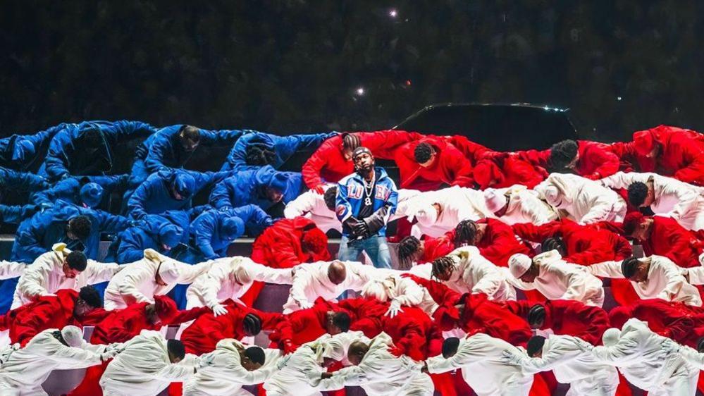 Rapper Kendrick Lamar standing with his arms crossed in the middle of a large group of dancers wearing red, white and blue in the formation of the US flag, during one of his gigs