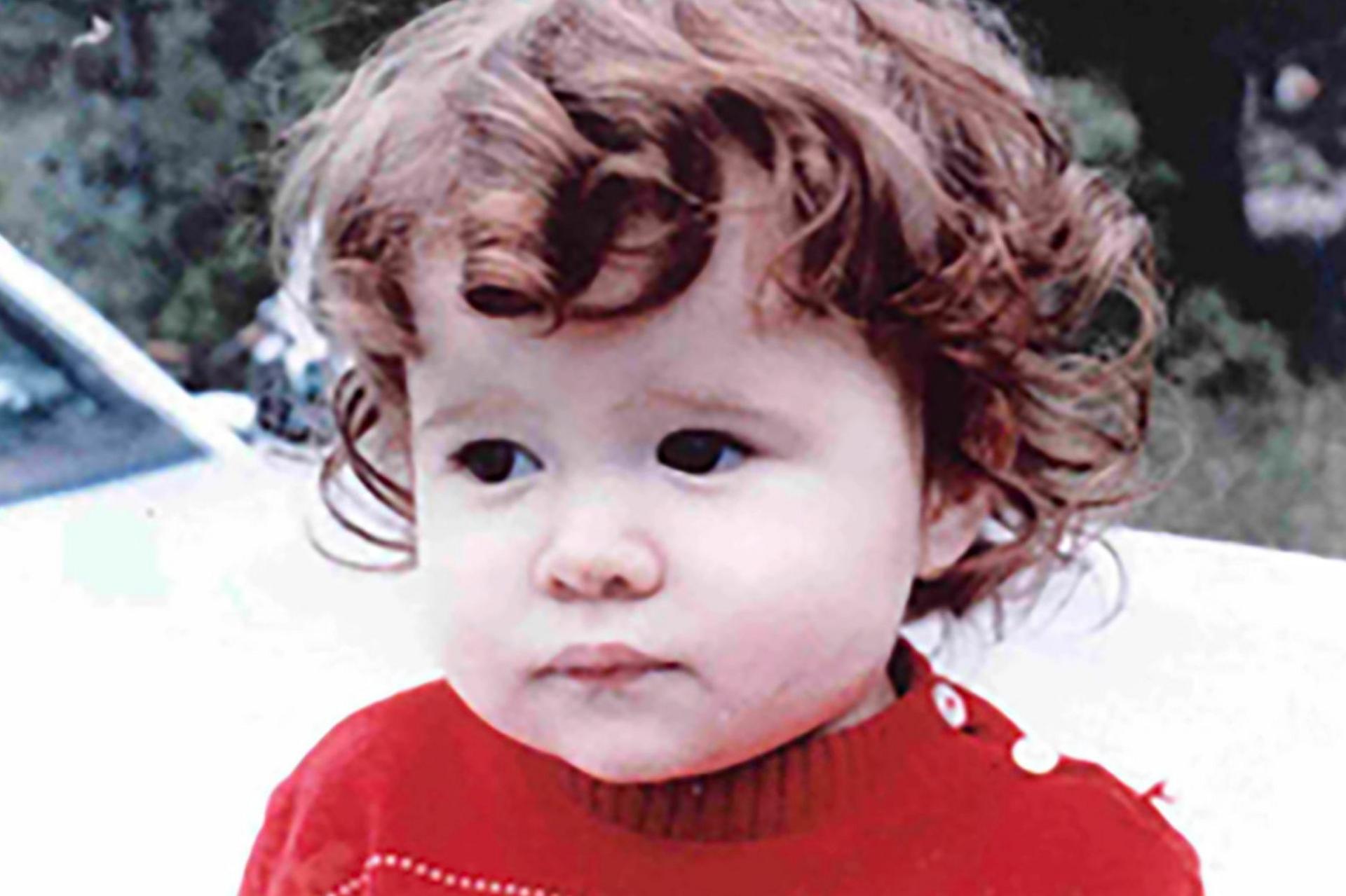 Christa Pike as a toddler. She has brown, short curly hair and is wearing a red jumper. The background appears to show part of a white vehicle