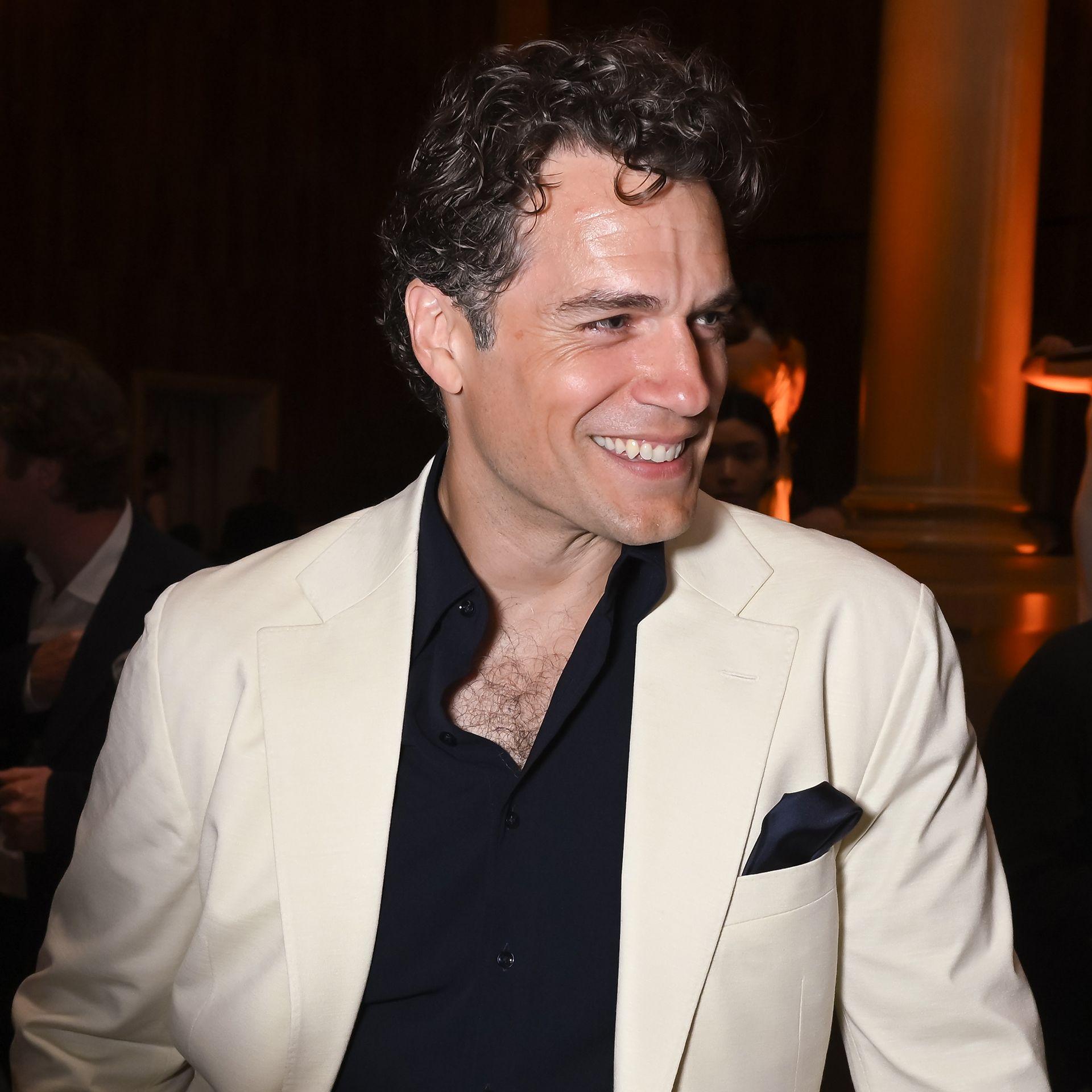 Henry Cavill smiles looking to right of frame while wearing a white suit jacket and black shirt, in London in June.