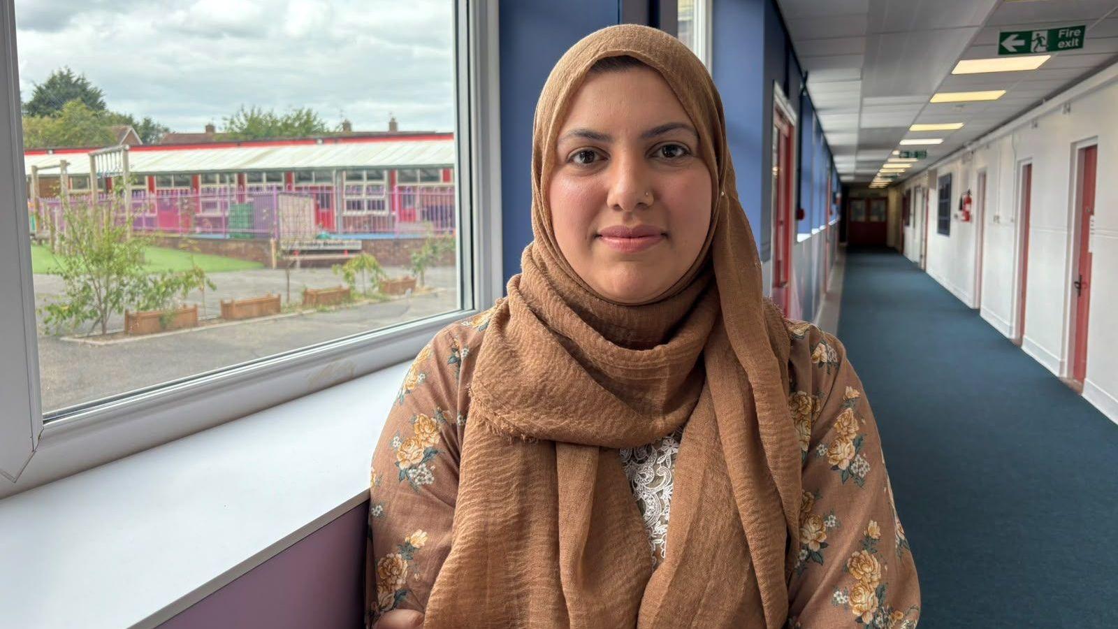 Mum Kinza is smiling at the camera. She is wearing a light brown top with flowers on, and light brown headscarf. She is standing in a school corridor and there is a playground behind her outside the window
