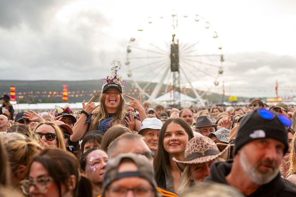 A crowd of people at a festival. One woman is sitting on someone's shoulders with lots of other people around her. She has an elaborate hair band over the top of a black baseball cap and is sticking her tongue out while gesturing with her hands